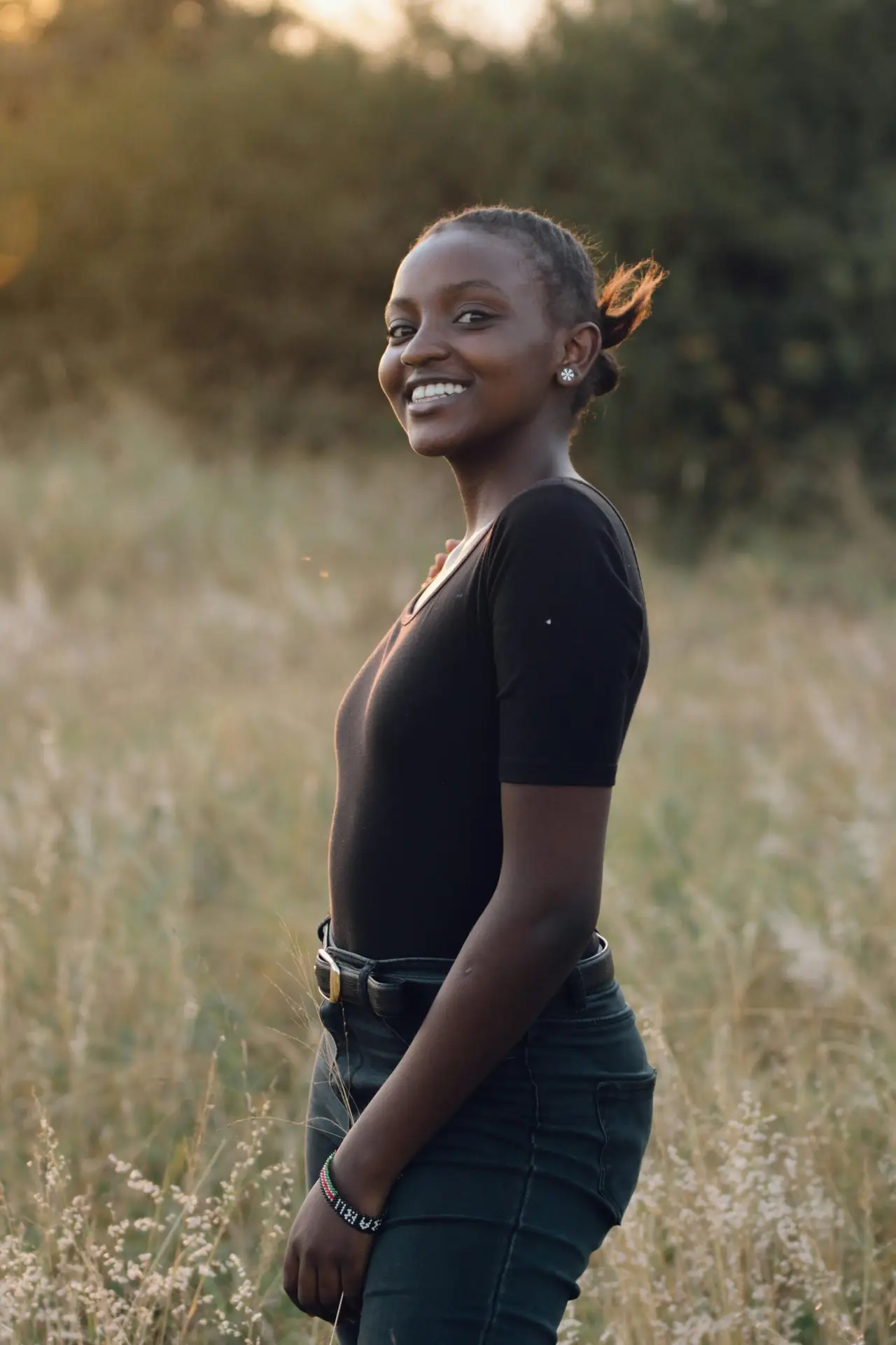 A smiling woman wearing a black top stands in a grassy field during sunset, embodying natural beauty and joy.